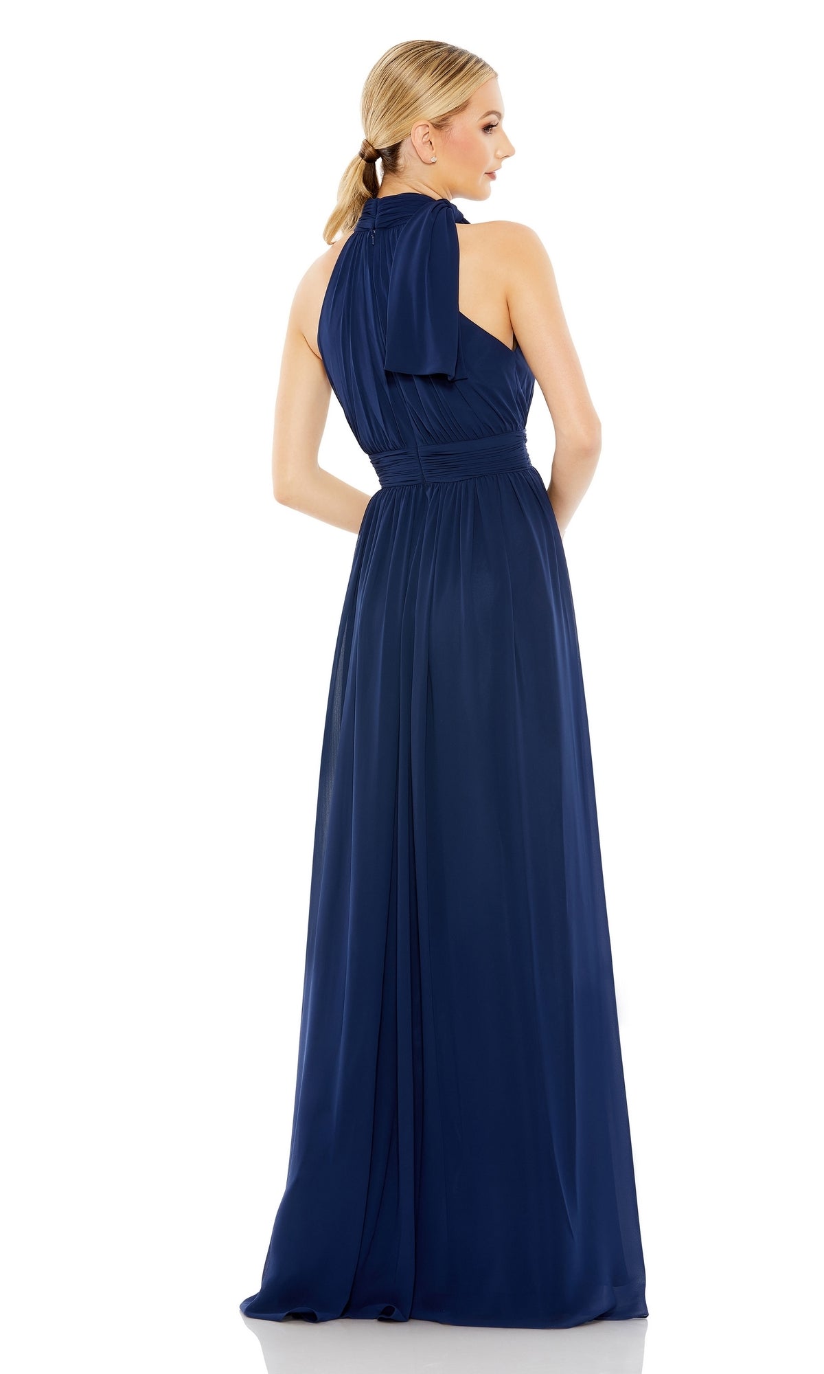 A woman models the Mac Duggal 55035 High-Neck Long Formal Dress, facing away to highlight the sleeveless design, floor-length navy blue fabric, back details, and tied halter neckline.